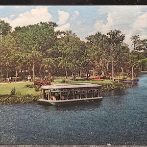 May include: Vintage postcard featuring a scenic river view with a passenger boat. Lush green trees line the riverbanks, and a blue sky with fluffy clouds fills the background. People are seen walking along the grassy shore.