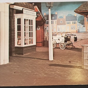 May include: A vintage postcard depicting a quaint colonial town square with a wooden building labeled "Cabinet Maker". A white horse-drawn cart with the text "Vanilla Ice" is parked in the square. The scene is painted in muted tones, giving it a nostalgic feel.