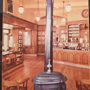 May include: A vintage interior with a wood-burning stove, wooden chairs, and hanging pendant lights. The space has a pharmacy or apothecary theme, with shelves and display cases filled with bottles and jars. The color scheme is warm, with shades of brown and beige.