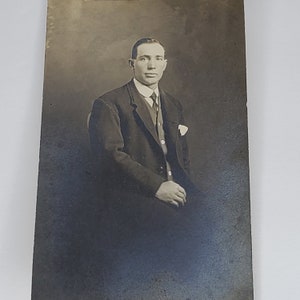 Puede incluir: Una fotografía en blanco y negro de un hombre con traje y corbata. Está sentado en una silla y mirando directamente a la cámara.