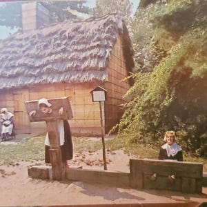 Puede incluir: Una mujer con un vestido blanco y un sombrero negro se encuentra en un cepo de madera. Dos niños con ropa similar se encuentran detrás de ella. Un edificio de madera con techo de paja se encuentra en el fondo.