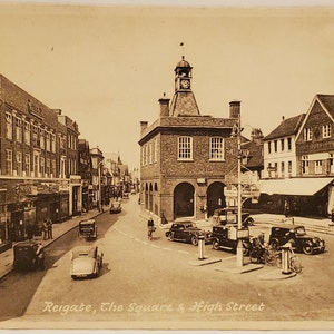 May include: A black and white vintage postcard showing a view of Reigate, The Square & High Street. The image shows a street scene with a clock tower building in the center, shops and buildings on either side, and cars parked on the street.