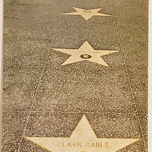 May include: A close-up view of a sidewalk with five stars embedded in the concrete. The stars are all the same size and shape, and they are all made of a light-colored material. The stars are arranged in a line, and they are all facing the same direction. The stars have the names of famous people inscribed on them, including "Clark Gable" and "Richard Dix".