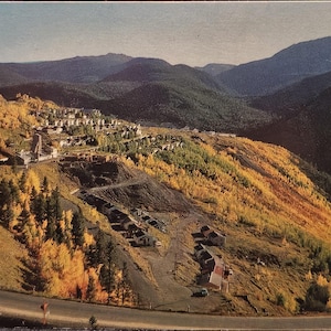 May include: A vintage postcard depicting a mountain landscape with a winding road. The hillside is covered in trees with yellow and green foliage. A small town is nestled on the hillside, with mountains in the background under a blue sky.