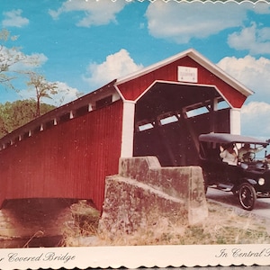 Op de afbeelding: Een rode houten overdekte brug met witte versieringen en een zwart interieur. Een oldtimer rijdt uit de brug. De brug is gelabeld "Sam Wagner Covered Bridge" en de tekst "In Central Pennsylvania" staat onderaan de afbeelding.