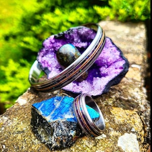 Puede incluir: Dos anillos de plata con diseños de incrustaciones de color púrpura y cobre. Los anillos están sobre una roca de cristal púrpura y blanca y una roca de cristal azul.