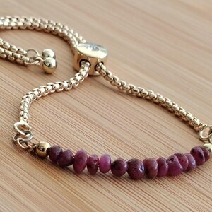 Pode incluir: Um bracelete de corrente dourada com fecho deslizante e uma fila de pequenas contas de pedras preciosas vermelho-escuras.