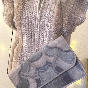 May include: A light gray clutch with a flap closure and a long, thin strap. The clutch features a patchwork design with shades of gray and blue. A beige, textured vest is in the background.