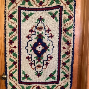 May include: A rectangular, off-white rug with a detailed floral design. The rug features a central pattern in blue, red, and brown, surrounded by a red border and a blue outer edge. Green leaves and red flower accents are spread throughout the design.