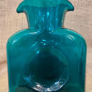 May include: Teal-colored glass bottle with a unique, rounded shape. The bottle has a wide, flat base and a narrow neck with a flared top. A circular indentation is visible on the front. The glass has a glossy finish, reflecting light.