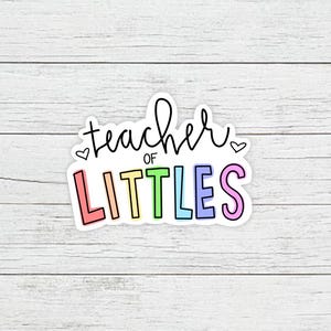 May include: A sticker with the text "teacher of littles" in rainbow colors with hearts on either side of the text.