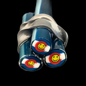 May include: A cluster of three blue glass rods, each with a Colorado flag design and a yellow smiley face. The rods are held together by a silver metal band, set against a black background.