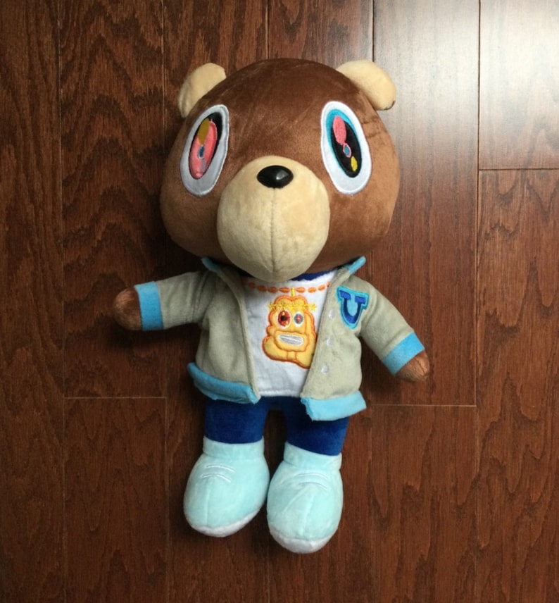 Pu&ograve; includere: Un orso di peluche marrone che indossa una giacca grigia, jeans blu e scarpe da ginnastica blu e bianche. L'orso ha grandi occhi colorati e una maglietta gialla con un viso di cartone animato.