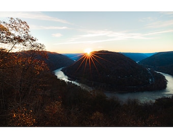 Könnte beinhalten: Panoramablick auf einen Fluss, der sich durch ein Tal schlängelt, mit einer leuchtend orangefarbenen Sonne, die hinter einem bewaldeten Hügel untergeht.
