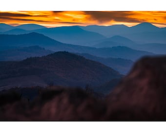 Könnte beinhalten: Ein Panoramablick auf eine Bergkette bei Sonnenuntergang. Der Himmel ist leuchtend orange und gelb, und die Berge sind tiefblau. Der Vordergrund ist eine Nahaufnahme eines felsigen Aufschlusses.