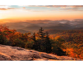 May include: A panoramic view of a mountain range with a rocky foreground. The mountains are covered in trees with fall foliage in shades of red, orange, and yellow. The sky is a soft orange and pink, indicating a sunrise or sunset.