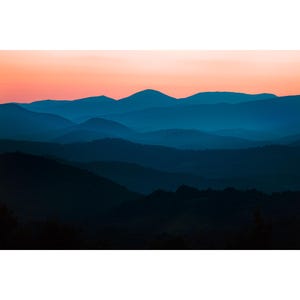 以下が含まれることがあります： 夕暮れの山脈のパノラマビュー。空は柔らかなオレンジとピンクで、山は濃い青色です。山は重なり合っており、最も近い山は最も暗く見えます。