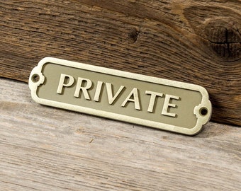May include: A gold-colored metal sign with the word "PRIVATE" embossed on it. The sign is on a wooden surface.