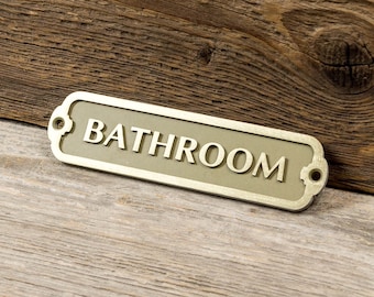 May include: A gold-colored metal sign with the word "BATHROOM" on it. The sign is on a wooden surface.