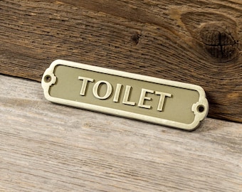May include: A gold-colored metal sign with rounded edges and the word "TOILET" in raised lettering.