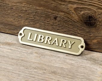 May include: A gold-colored metal sign with rounded edges and a raised inscription that reads "LIBRARY". The sign is on a wooden surface.