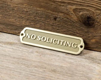May include: A rectangular metal sign with rounded ends and a light olive green center. The sign features the text NO SOLICITING in raised, light-colored capital letters. It includes two small circular holes on the left and right sides for mounting.