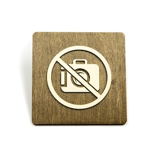 May include: A brown wooden sign with a white circle and a diagonal line through it. Inside the circle is a white silhouette of a camera. The sign says "No Photography".