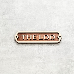 以下が含まれることがあります： 白い文字で「THE LOO」と書かれた、金色の縁取りのある木製の看板。灰色の壁に取り付けられています。