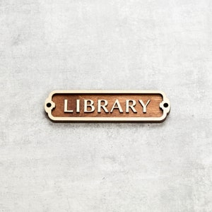 Peut inclure: Une pancarte en bois aux bords arrondis et à la finition brun clair. La pancarte indique "LIBRARY" en lettres blanches.