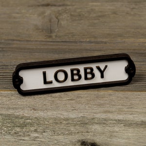 Peut inclure: Une pancarte noire et blanche avec le mot "LOBBY" en lettres dorées. La pancarte est en bois et a un bord arrondi.