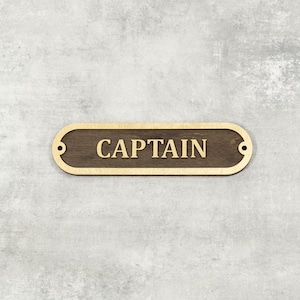 May include: Wooden sign reading "CAPTAIN" in gold lettering. The sign is a dark brown oval shape with a gold border and two small holes at each end. It is mounted on a light grey textured wall.
