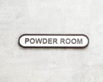 May include: A white oval sign with a black border and the text "POWDER ROOM" in black lettering.