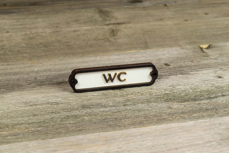 Peut inclure: Une plaque rectangulaire blanche avec une bordure noire et des coins arrondis. La plaque indique "WC" en lettres dor&eacute;es.