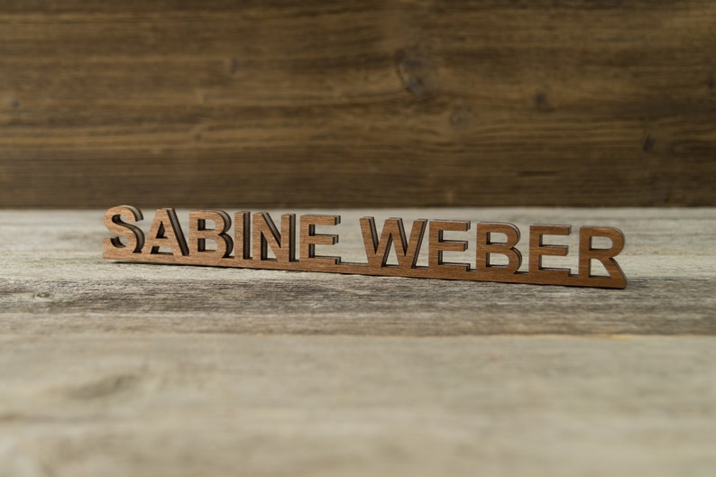 May include: Wooden name sign with the name "SABINE WEBER" in brown letters on a light brown wooden background.