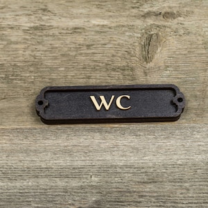 Peut inclure: Une plaque rectangulaire noire avec des coins arrondis et un lettrage dor&eacute; qui indique "WC" sur un fond en bois.