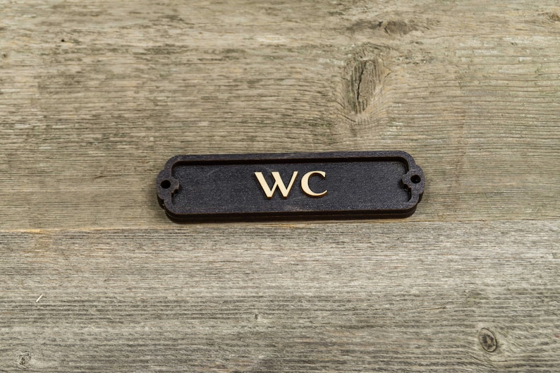 Peut inclure: Une plaque rectangulaire noire avec des coins arrondis et un lettrage dor&eacute; qui indique "WC" sur un fond en bois.