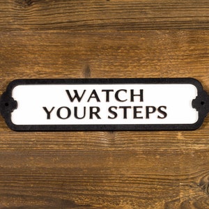 Op de afbeelding: Een zwart-wit bord met de tekst "WATCH YOUR STEPS" op een houten achtergrond.