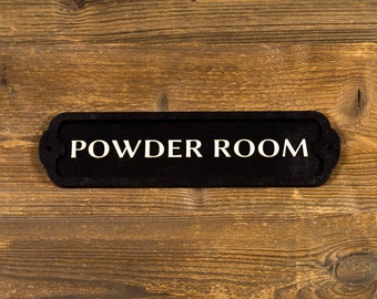 May include: Black sign with white lettering that reads "POWDER ROOM" on a wooden background.