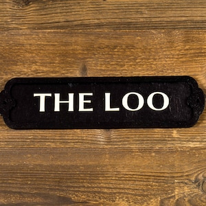 May include: Black wooden sign with white lettering that reads "THE LOO" on a brown wooden background.