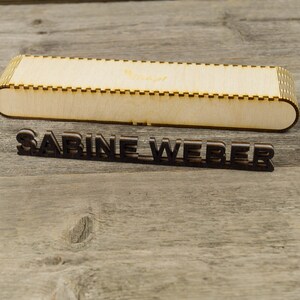 May include: A wooden box with the name "SABINE WEBER" engraved on it. The box is made of light wood and has a simple design.