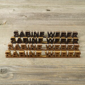May include: Four wooden name plaques stacked on top of each other, each with the name "Sabine Weber" in black and gold letters.