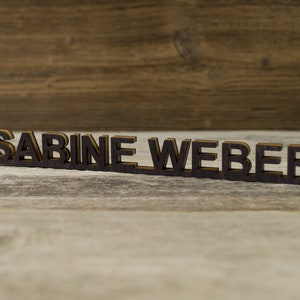 May include: A wooden sign with the name "SABINE WEBER" spelled out in black letters.