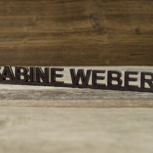 May include: A wooden nameplate with the name "SABINE WEBER" spelled out in black letters.