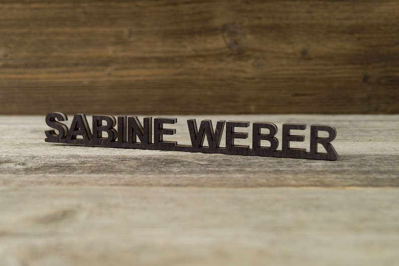 May include: A wooden nameplate with the name "SABINE WEBER" spelled out in black letters.