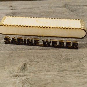 May include: A wooden box with a cutout design and the name "SABINE WEBER" written in black letters on a wooden surface.