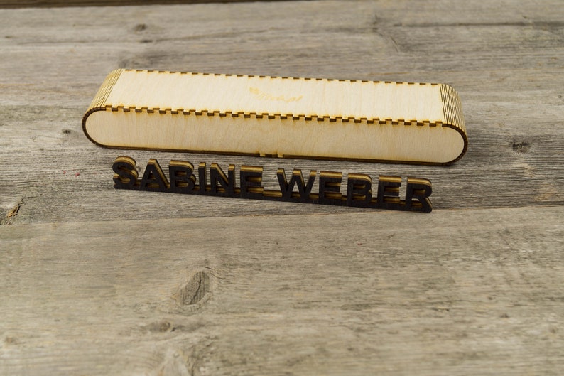 May include: A wooden box with a cutout design and the name "SABINE WEBER" written in black letters on a wooden surface.