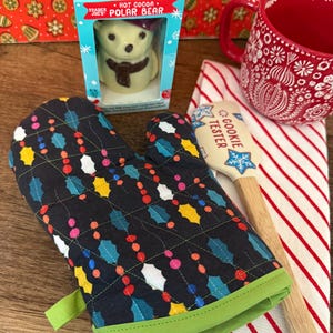 May include: A black and green oven mitt with a quilted pattern of holly leaves and colorful lights. A wooden cookie tester with the text "Cookie Tester" is on a red and white striped kitchen towel. A red mug with a white snowflake design and a box of Trader Joe's hot cocoa polar bear are in the background.