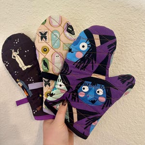 May include: Three quilted oven mitts with Halloween-themed designs. One mitt features a witch with a purple and black design, another has a ghost and a cat with a pink and gold design, and the third has a woman with stars and a purple and black design.