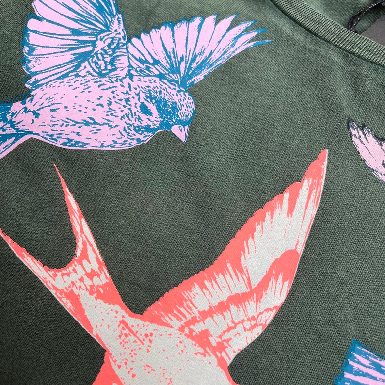 May include: A green t-shirt with a pink and blue bird and a pink and white bird printed on it.