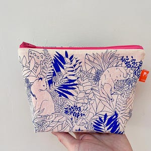 Peut inclure: Petite trousse de maquillage rectangulaire avec fermeture éclair rose. Le sac présente un fond rose clair avec un imprimé botanique et animalier bleu et blanc. Le sac est tenu par une main.